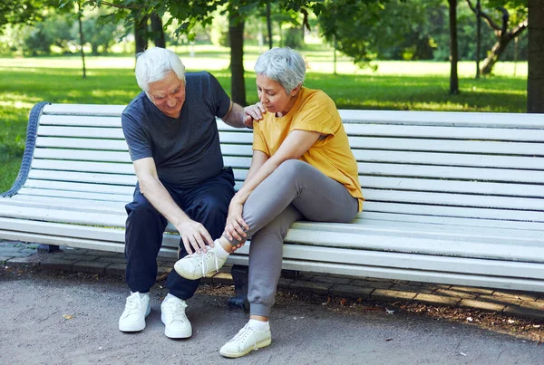 During morning stroll, sportive exercises in park, elderly 60s woman got injured ankle, grip foot sit in bench with caring disappointed husband. Traumas, injures of older people concept