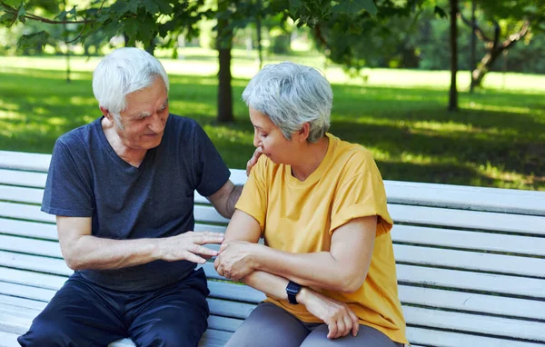 During morning sportive stroll, sportive exercises in park, elderly 60s woman got injured shoulder gripping arm sit in bench with caring disappointed husband. Traumas, injures of older people concept