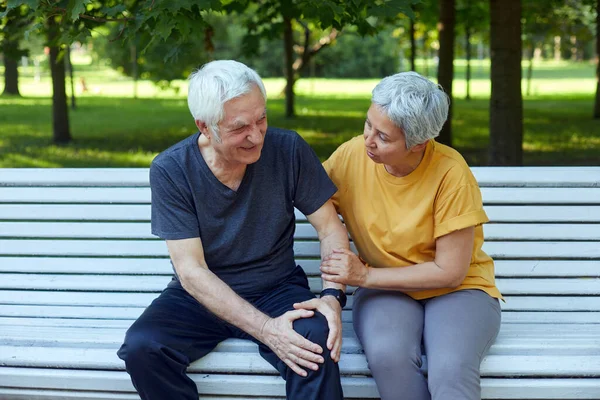 During morning sportive stroll or making exercises in a park, elderly 60s man got injured his knee, gripping leg sit in bench with caring disappointed wife. Traumas, injures of older people concept