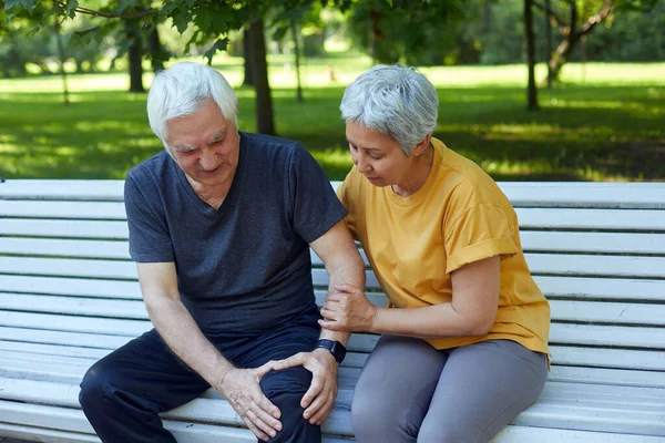 During morning sportive stroll or making exercises in a park, elderly 60s man got injured his knee, gripping leg sit in bench with caring disappointed wife. Traumas, injures of older people concept