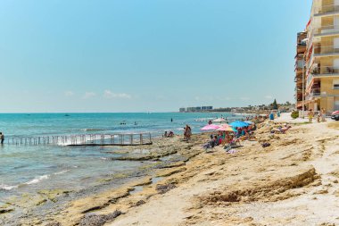 Torrevieja, Spain - August 2, 2022: Tourists sunbathing on rocky beach, enjoy Mediterranean Sea spend vacation on sandy coastline. Summer holidays. Costa Blanca, Province of Alicante. Spain