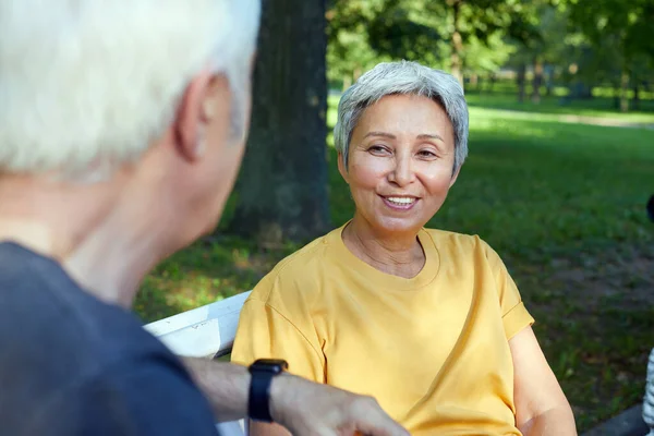 Attractive older couple talking seated on bench in summer park, wear sportive clothes, enjoy conversation in the morning outdoors. Communication, harmonic marriage, unity concept