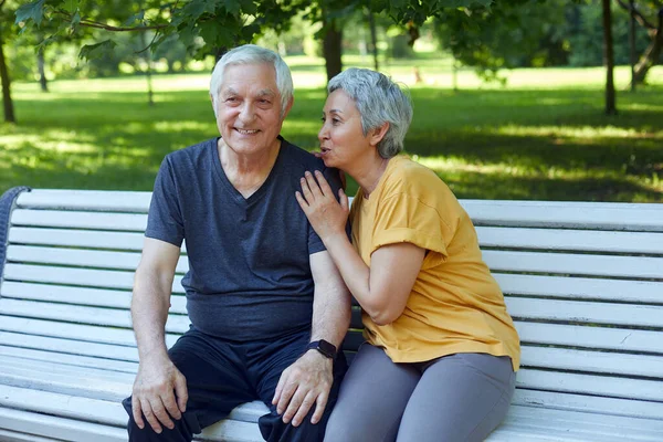 Attractive older couple talking seated on bench in summer park, wear sportive clothes, enjoy conversation in the morning outdoors. Communication, harmonic marriage, unity, rumours concept