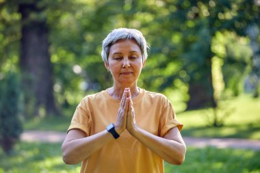 Yaşlı, aktif Asyalı kadın yaz parkında gözleri kapalı meditasyon pratiği yapıyor. Kollarını kavuşturmuş, Mudra veya Namaste işareti yapıyor. Yoga yapıyor, dışarıda yoga yapıyor.