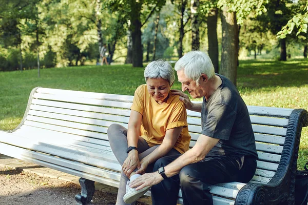 Elderly 60s woman got injured her ankle during morning stroll in summer park, grip foot seated in bench with caring disappointed husband. Traumas, injures of older people, medical insurance concept