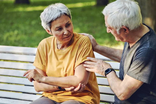 During morning sportive stroll or making exercises in park, old 60s woman got injured her shoulder or elbow gripping arm sit in bench with caring disappointed husband. Traumas of older people concept