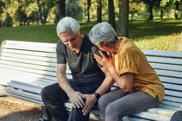 During morning sportive stroll in summer park, elderly 70s man got injured his knee, gripping leg seated in bench with caring disappointed wife. Physical traumas of older people. Medicine concept