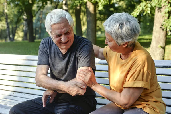 During morning sportive stroll or making exercises in park, elderly 70s man got injured his shoulder or elbow gripping arm sit in bench with caring disappointed wife. Traumas of older people concept