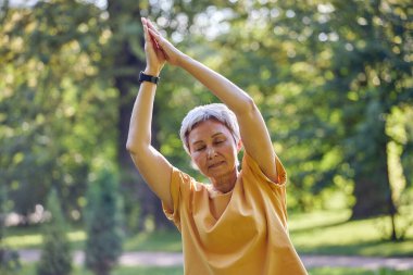 Spor giyim sektöründe aktif olan yaşlı bir Asyalı kadın yaz parkında avuçlarını kıvırarak meditasyon yaparken esneme egzersizleri yapıyor, dışarıda yoga yapıyor. Emeklilikte sağlıklı yaşam tarzı