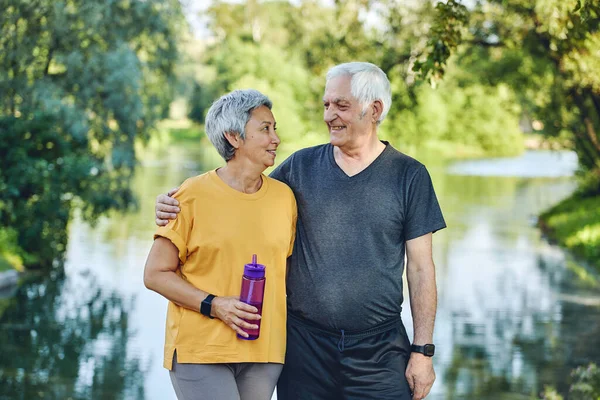 Attractive older couple in sportswear hugging pose look at each other standing in summer park near lake after morning stroll, sport active lifestyle holds bottle with still water. Healthy retired life