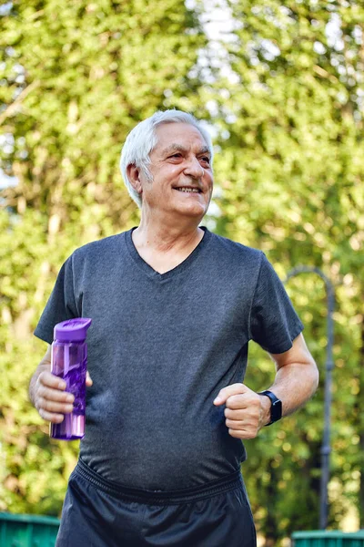 Older man in sportswear holds reusable plastic bottle standing or jogging in summer park, caring about health, enjoy morning work out. Healthy lifestyle
