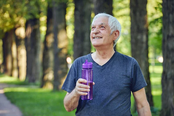 Older man in sportswear holds reusable plastic bottle standing or jogging in summer park, caring about health, enjoy morning work out feels happy. Healthy sportive lifestyle of retirees concept