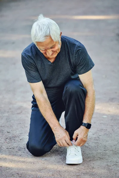 Elderly Caucasian man in sportive wear ties shoelaces ready for morning jogging or sportive walk stroll in summer park. Healthy active lifestyle of retired person, work out concept