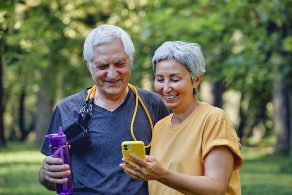 Attractive elderly grey-haired wife and husband using modern smartphone check calories counter or tracking after sportive work out in summer park. Tech usage for sport and healthy lifestyle