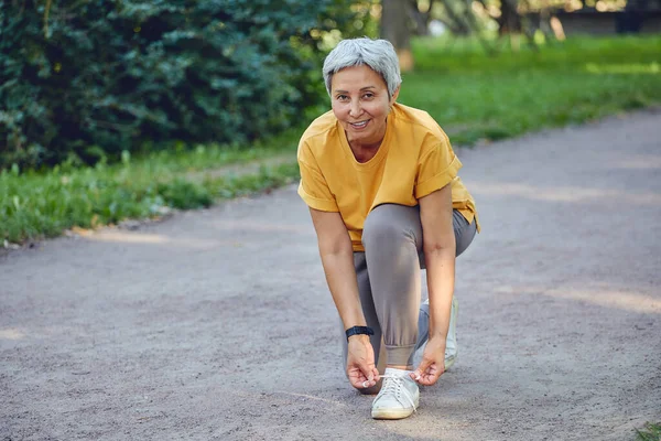Elderly middle-aged Asia woman in sportive wear ties shoelaces ready for morning jogging or sportive walk stroll in summer park smile look at camera. Healthy active lifestyle of retiree, work out