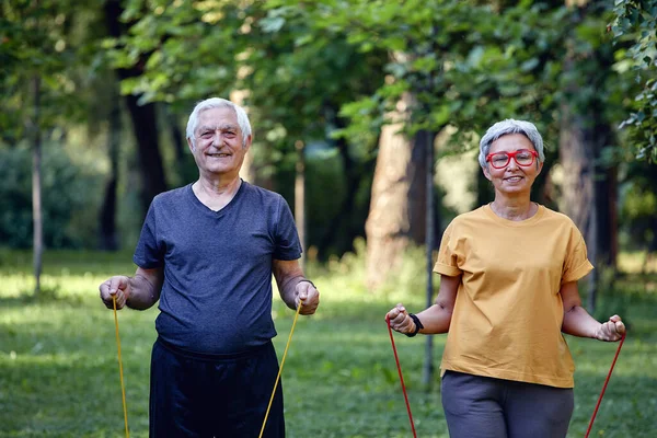 シニア配偶者は 抵抗ゴムバンドを使用して午前中に夏の公園の屋外でスポーツウェアを着用します 健康的なライフスタイル アクティブな退職生活は近代的な家電コンセプトを使用します — ストック写真