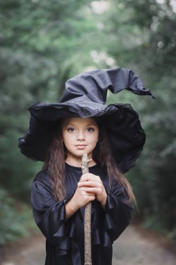 Cadılar Bayramı konsepti olan, ağacın yanındaki ormanda cadı kostümü giymiş küçük bir kız.
