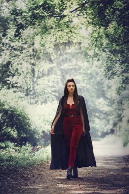 Siyah pelerinli inanılmaz güzel bir cadı sık ormanda yürüyor. Cadılar Bayramı konsepti