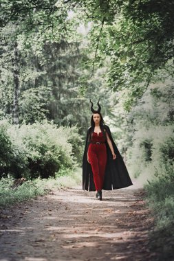 Kara pelerinli, boynuzlu inanılmaz güzel bir cadı sık ormanda yürüyor. Cadılar Bayramı konsepti