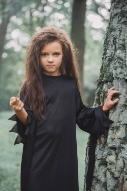 Cadılar Bayramı konsepti olan, ağacın yanındaki ormanda cadı kostümü giymiş küçük bir kız.