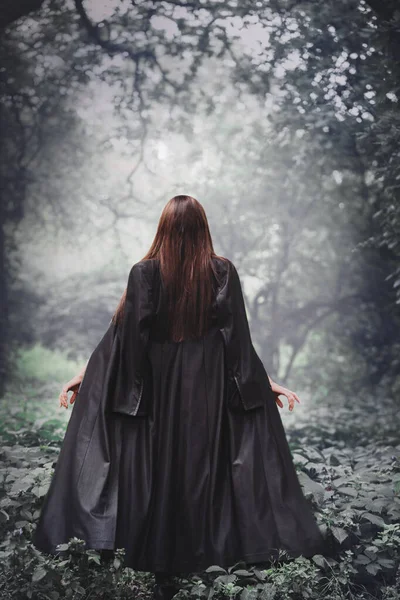 A young woman in a black raincoat walks in the dark of the forest, back view. Halloween concept