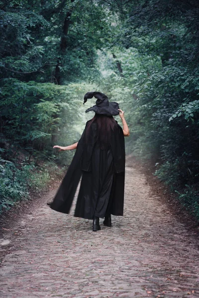 A witch in a black cloak and a big black hat in a dark forest. Halloween concept