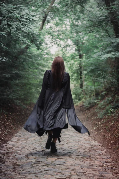 黒いレインコートを着た背の高い美しい女性が鬱蒼とした森の中を歩いています ハロウィンのコンセプト — ストック写真