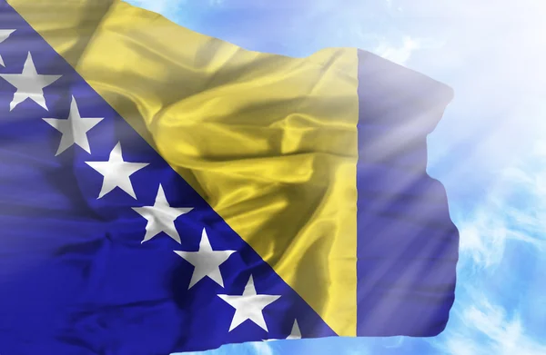 Bosnien und Herzegowina schwenkt Flagge gegen blauen Himmel mit Sonnenstrahlen — Stockfoto