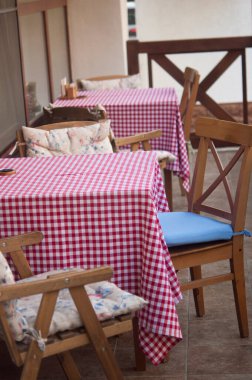 Ağaçların ve çiçeklerin arasındaki tuğla duvarın yanında ekose masa örtüsü olan boş masalar.