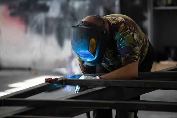 Разнорабочий выполняет сварку и шлифование на своем рабочем месте в мастерской, в то время как искры "летают" вокруг него. На нем защитный шлем и снаряжение..