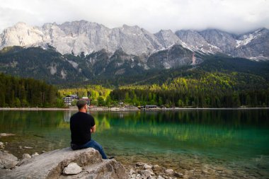 Bir kayanın üzerinde oturan ve Bavyera Alplerinde Eibsee Gölü üzerinde güzel bir manzara izleyen bir turist. Avrupa 'nın ünlü turistik beldesi