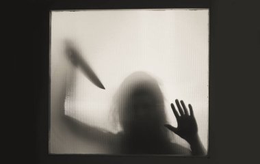 Korku, cadılar bayramı arkaplanı, camın arkasında elinde bıçak tutan karanlık bir figür.