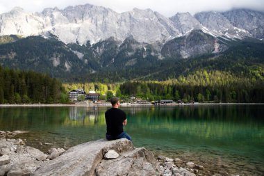 Bir kayanın üzerinde oturan ve Bavyera Alplerinde Eibsee Gölü üzerinde güzel bir manzara izleyen bir turist. Avrupa 'nın ünlü turistik beldesi