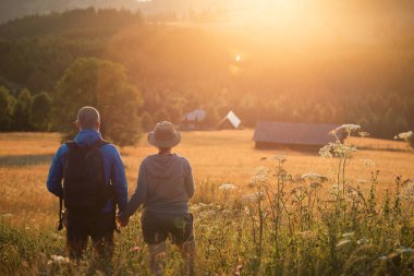 Hiker couple in summer meadow looking at magic sunset over hills. Relaxation, nature and tourism concept.