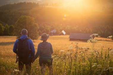 Hiker couple in summer meadow looking at magic sunset over hills. Relaxation, nature and tourism concept.