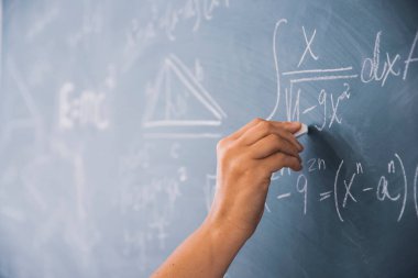 Tebeşiri tutan ve tahtaya matematiksel denklemler yazan bir elin yakın plan çekimi. Eğitim kavramı.