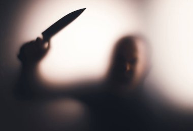 Korku, cadılar bayramı arkaplanı, camın arkasında elinde bıçak tutan karanlık bir figür.