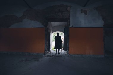 A mysterious figure wearing a long coat and fedora standing in a ruined, abandoned house