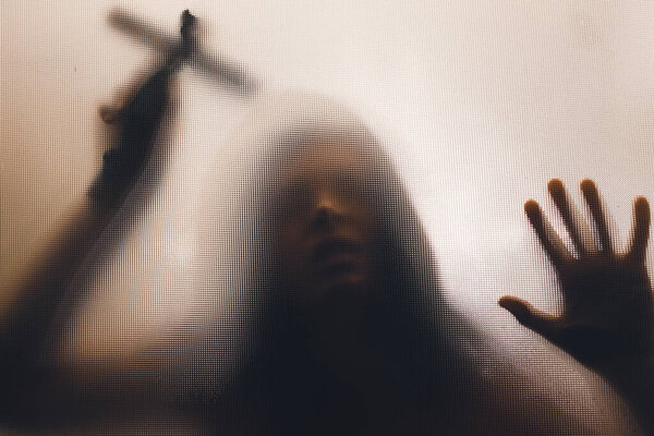 Horror ghost woman holding a cross behind the matte glass. Halloween festival concept.