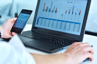 Yönetici ofisinde oturuyor ve ekranda stok verileri olan akıllı telefon ve dizüstü bilgisayar kullanıyor. İş ve iş kavramı. Kapat.