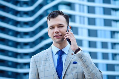 Happy young businessman in suit talking on mobile phone on skyscraper background. Business corporate and building concept.