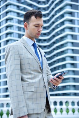 Takım elbiseli genç işadamı modern gökdelen arka planında cep telefonu kullanıyor. İş ve teknoloji kavramı. Kapat.