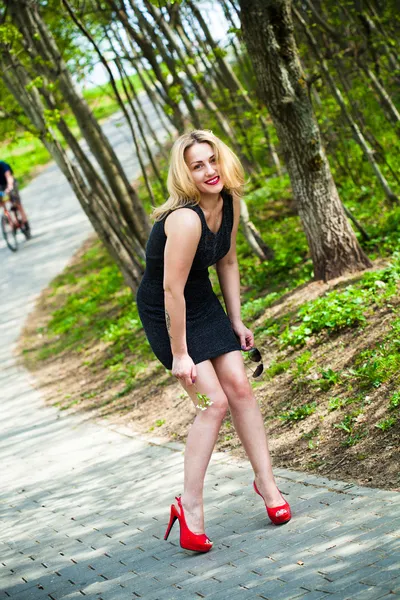 Porträtt av en leende vacker flicka i naturen. i en svart klänning och röda skor. — Stockfoto