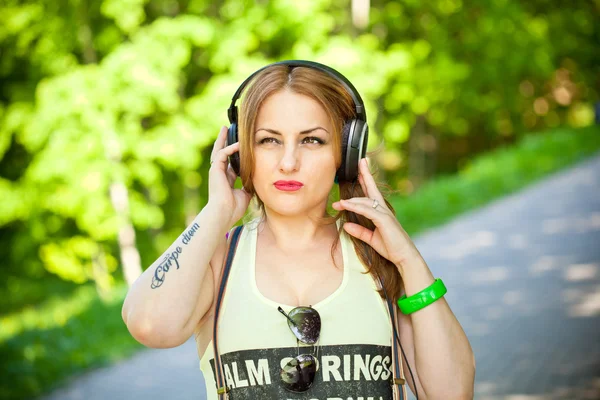 Beautiful young girl in a T-shirt intently listening to music with headphones outdoors — Stock Photo, Image