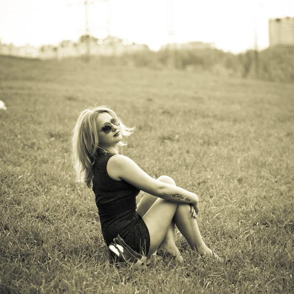 Beautiful young blond woman sitting on the grass. In sunglasses, a black dress, barefoot. Sepia toned photo — Stock Photo, Image