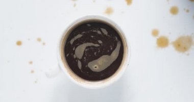 Bir fincan kahvenin içindeki köpük, üzerinde kahve damlaları olan beyaz bir masada saat yönünde dönüyor. Yukarıdan görüntüle.