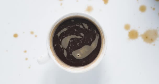 コーヒーのコップの泡はコーヒーの低下と白いテーブルで時計回りに回転している 上からの眺め — ストック動画
