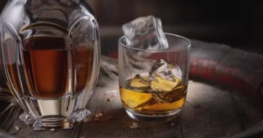 Bir buz küpü yavaşça viski bardağının üstüne dökülüyor. Eski bir viski fıçısının üstüne. İçkili bir sürahi yakınlarda duruyor, koyu kahverengi bir arka plan.. 