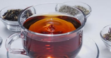Damla yavaşça bir bardak çaya düşer. Etrafta farklı yaprak çayları olan küçük kaseler var. Beyaz arkaplan. 