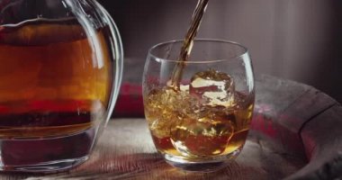 Viski fıskiyesi buz küpleriyle dolu bir bardağa dökülüyor, eski bir viski fıçısının üzerinde duruyor. İçkili bir sürahi yakında duruyor.. 